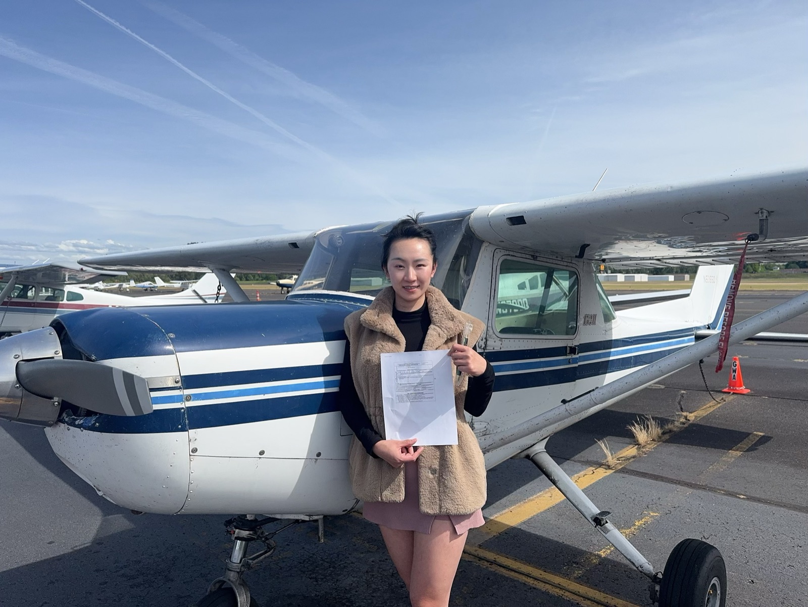 Nicole standing in front of a small Cessna aircraft on the tarmac, holding her pilot certificate paperwork after a training flight